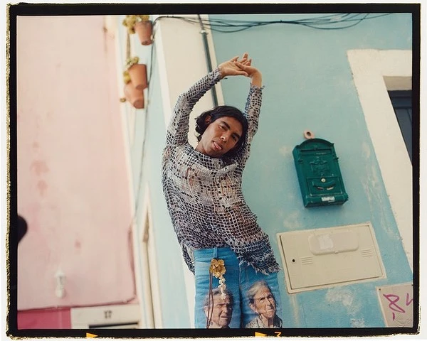 Man stretches against a light blue building on a street with pale pink buildings, showcasing artistic jeans and a patterned top in daylight.
