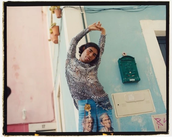Man stretches against a light blue building on a street with pale pink buildings, showcasing artistic jeans and a patterned top in daylight.