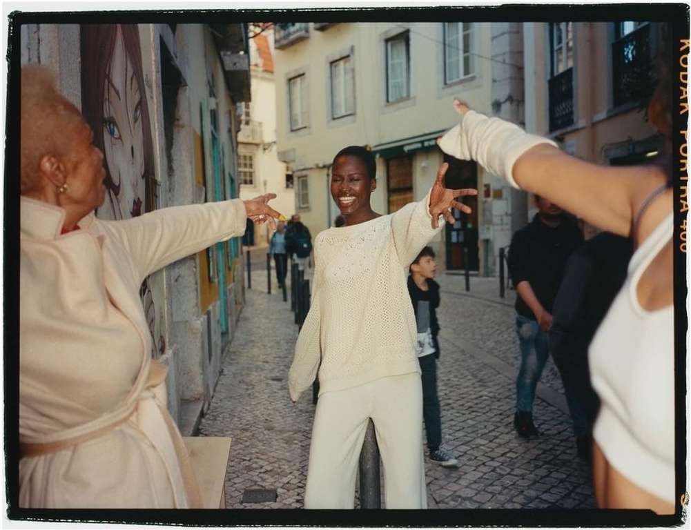 Three women in a cobblestone alleyway in cream-colored outfits, with others in the background, and buildings lining the alley.