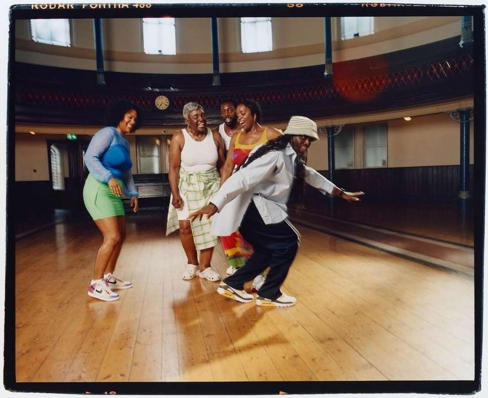 A diverse group of five people dancing indoors on a wooden floor in a well-lit, spacious hall with large windows and architectural details.
