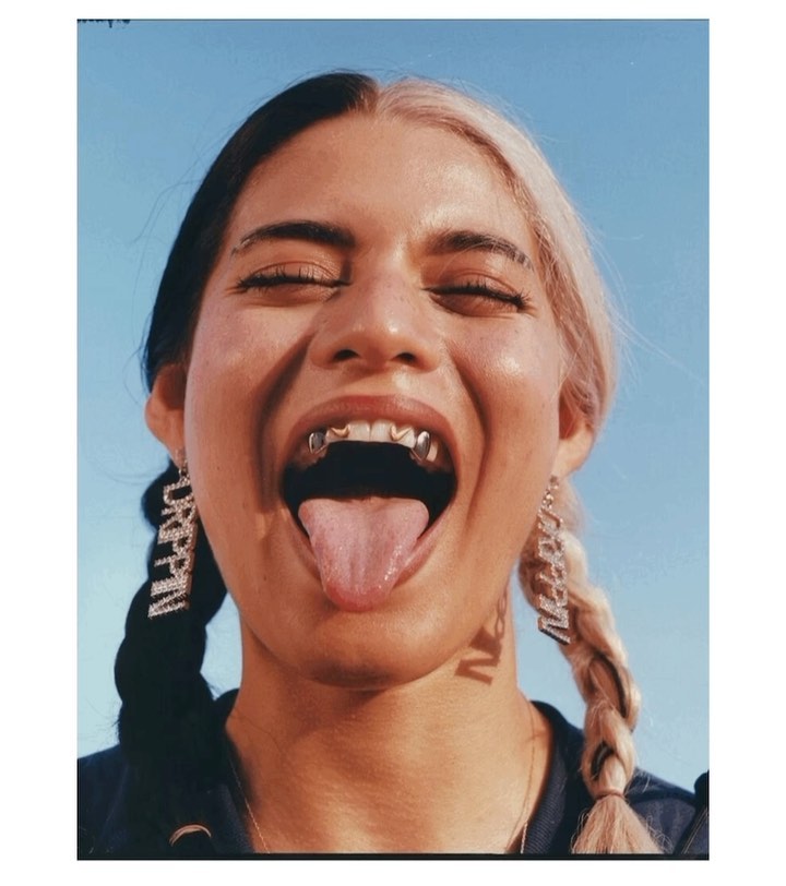 Close-up portrait of an adult woman with split-dyed hair, displaying gold fang grillz and tongue with blue sky in the background.