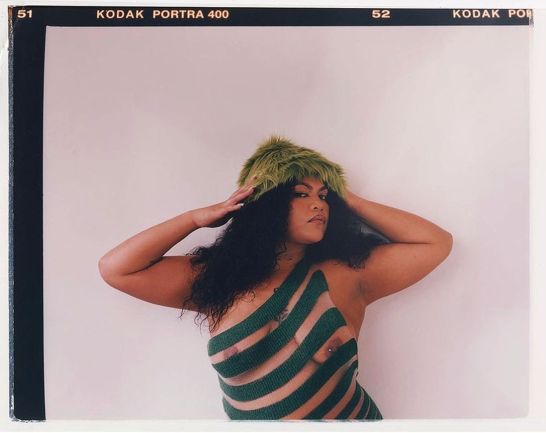 Portrait of a woman with dark curly hair wearing a striped green dress and a fuzzy green hat, striking a confident pose in studio.