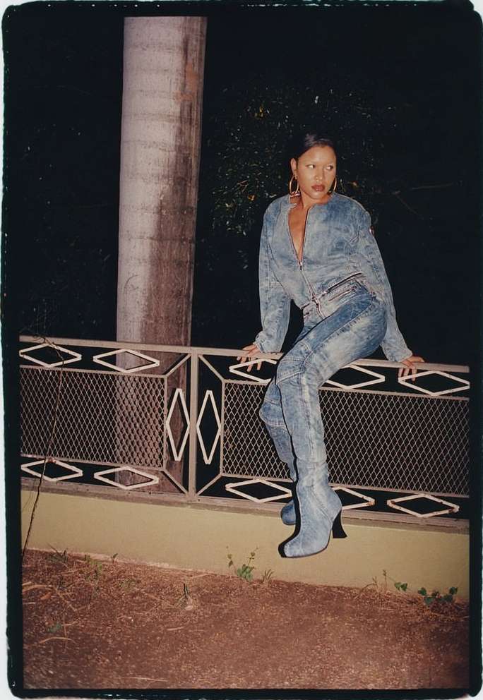 Woman in denim jacket and matching denim boots seated on an ornate railing at night outdoors
