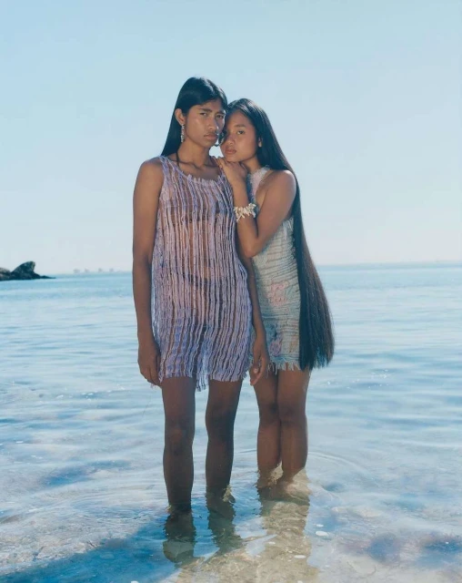 Two women in dresses stand in shallow water by the beach. The sunlight reflects in the water, creating a bright, summery scene.