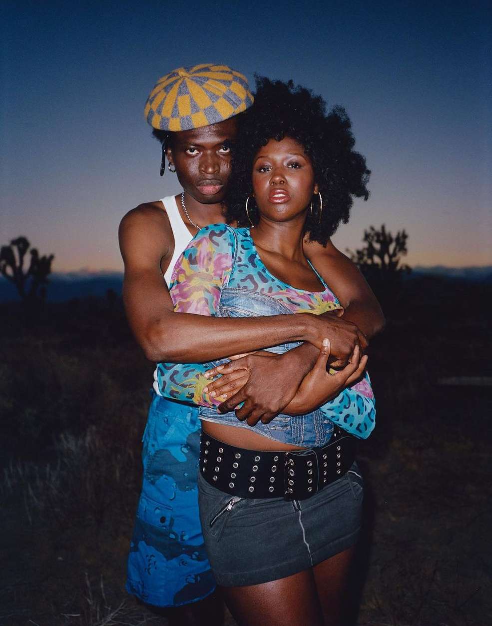 A stylish African-descent couple embraces in a desert setting at dusk, wearing fashionable clothing and posing intimately, captured in a medium shot.