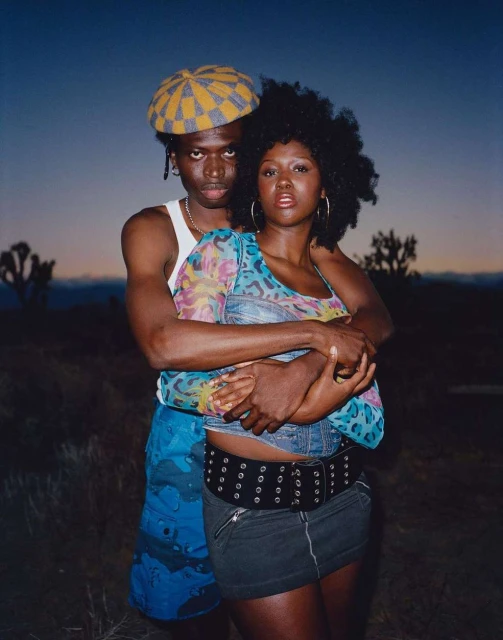 A stylish African-descent couple embraces in a desert setting at dusk, wearing fashionable clothing and posing intimately, captured in a medium shot.