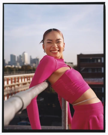 Smiling woman in hot pink athletic top leaning against a metal railing on a rooftop with a distant city skyline.