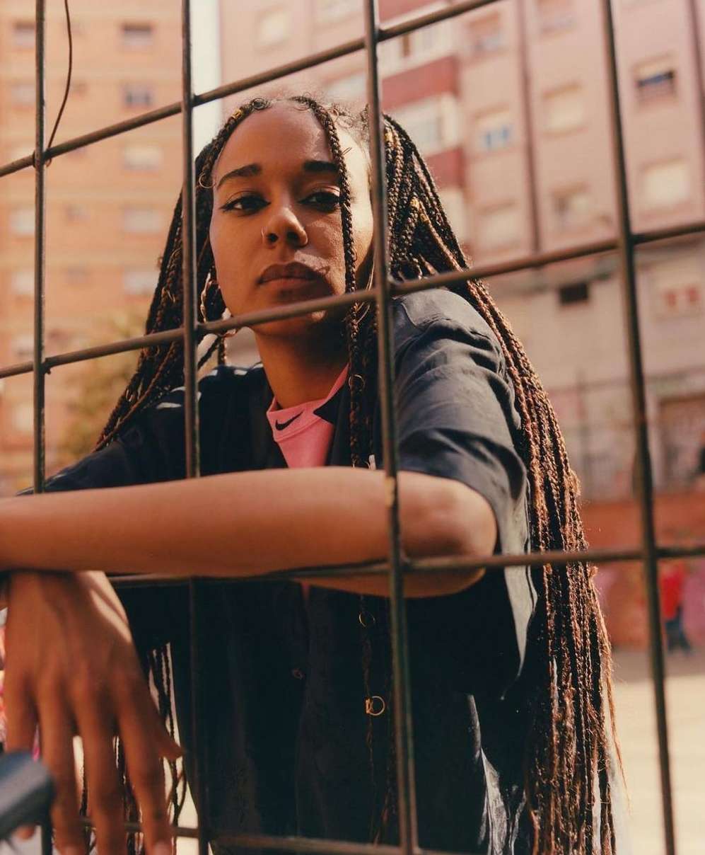 Portrait of a young adult woman with braids, leaning on a metal fence, wearing a pink top under a dark button-down shirt in an urban setting.