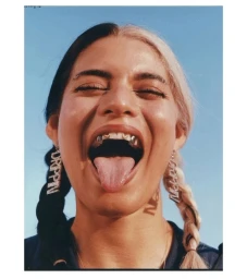 Close-up portrait of a woman with split-dyed black and blonde braided hair sticking her tongue out, wearing diamond drop earrings against a blue sky.