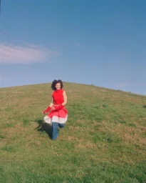 Smiling woman in red sleeveless top and blue jeans running down a grassy hill under a clear blue sky