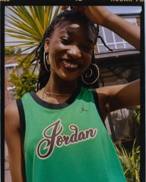 Smiling Black woman wearing a green mesh basketball jersey and large hoop earrings outdoors in London by Juanita Richards