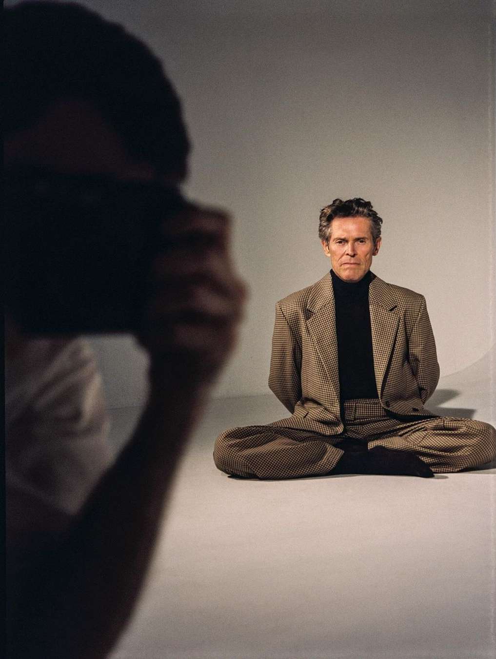 Actor Willem Dafoe photographed indoors sitting cross-legged, wearing a patterned brown suit, while a photographer is visible in the frame.