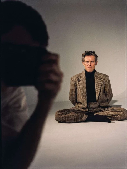 Actor Willem Dafoe photographed indoors sitting cross-legged, wearing a patterned brown suit, while a photographer is visible in the frame.