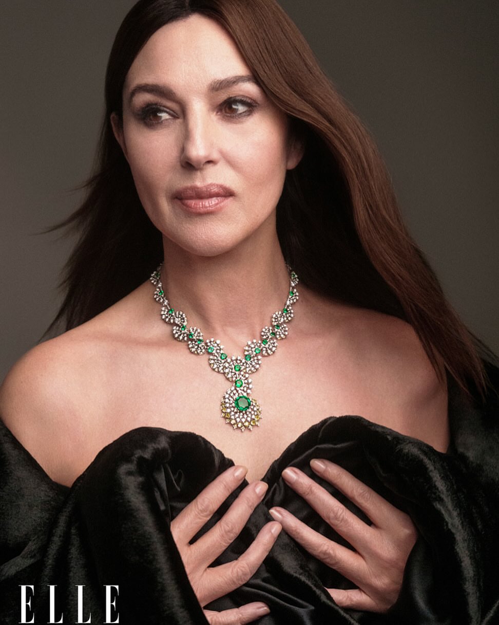 Portrait of Monica Bellucci wearing a floral emerald and diamond necklace, draped in a black velvet-like wrap, against a grey backdrop.