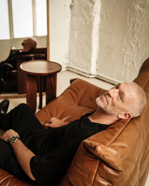 Man in black reclines on tan leather chair in minimalist room with side table and mirror reflecting same scene and light walls.
