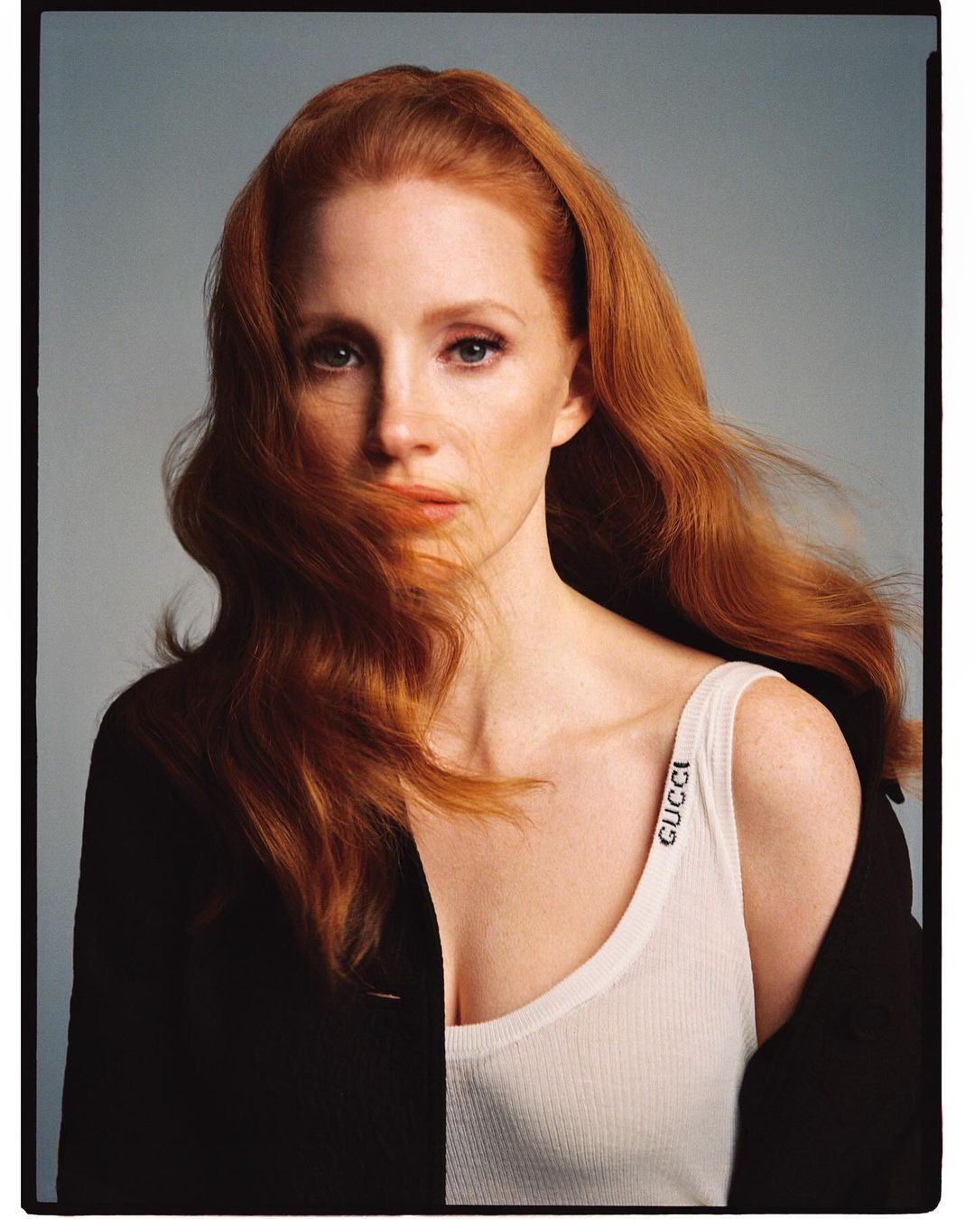 Close-up portrait of Jessica Chastain with red hair, wearing a white tank top with the word 'Gucci' on the strap and a black jacket.