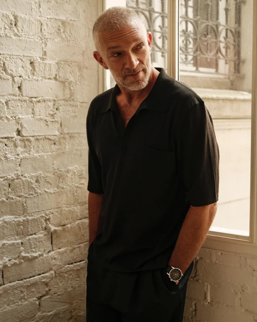 Portrait of Vincent Cassel standing near a window in dark clothing with a watch, captured in an indoor setting with neutral tones.