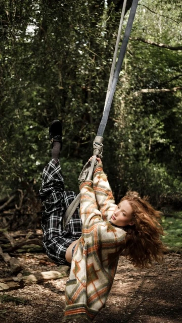 A woman with curly hair is swinging on a rope swing in a wooded area, wearing a patterned sweater and plaid pants, enjoying outdoor recreation.