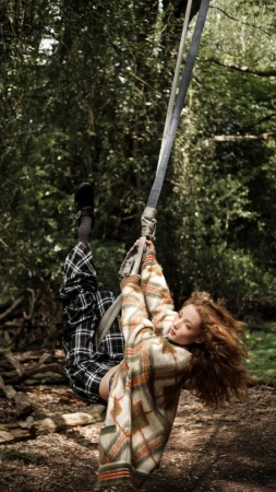 A woman with curly hair is swinging on a rope swing in a wooded area, wearing a patterned sweater and plaid pants, enjoying outdoor recreation.