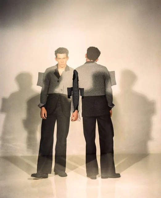 Two adult males wearing matching grey jackets and black pants face the camera and away, standing in a bright photo studio.