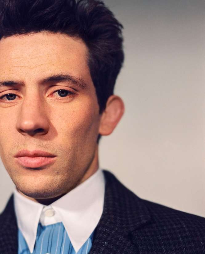 Close-up portrait of a light-skinned adult male with dark hair wearing a patterned suit jacket and blue striped shirt on a neutral background.