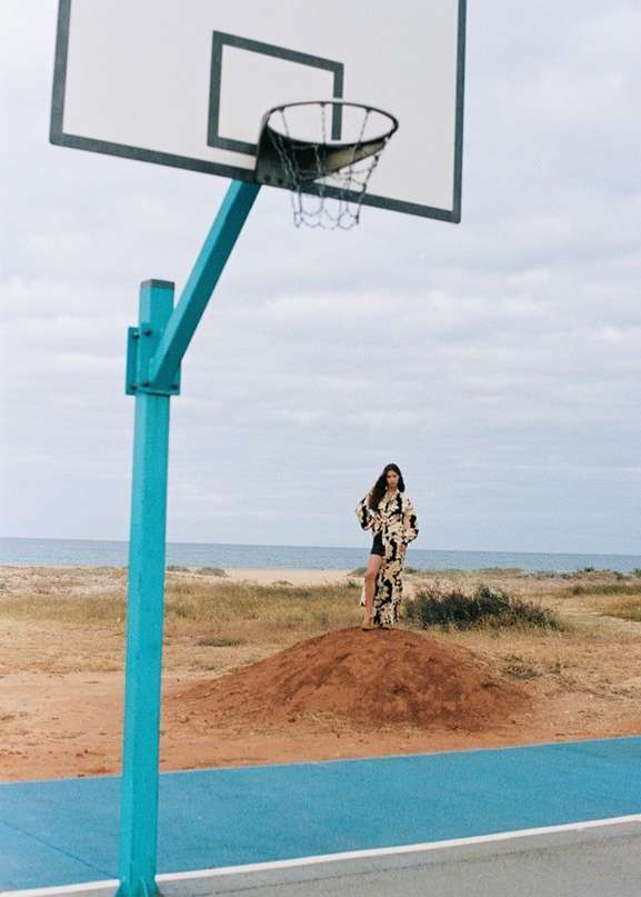 A lone female figure stands on a dune under a basketball hoop near the ocean, wearing a stylish, patterned outfit in a natural outdoor setting.