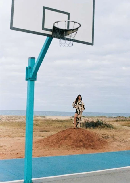 A lone female figure stands on a dune under a basketball hoop near the ocean, wearing a stylish, patterned outfit in a natural outdoor setting.