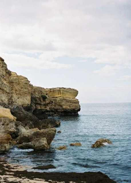 Coastal cliffs meet the ocean under a cloudy sky. Brown rock formations contrast with the blue water in this scenic landscape.