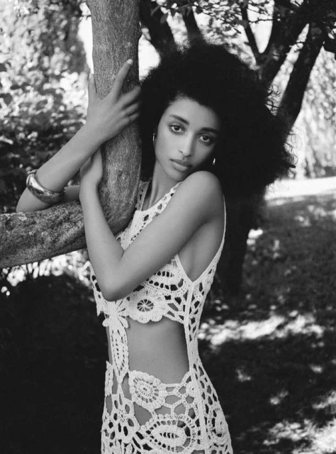 Black and white portrait of an adult woman with curly hair, wearing a crochet dress, posing against a tree in a natural outdoor setting.