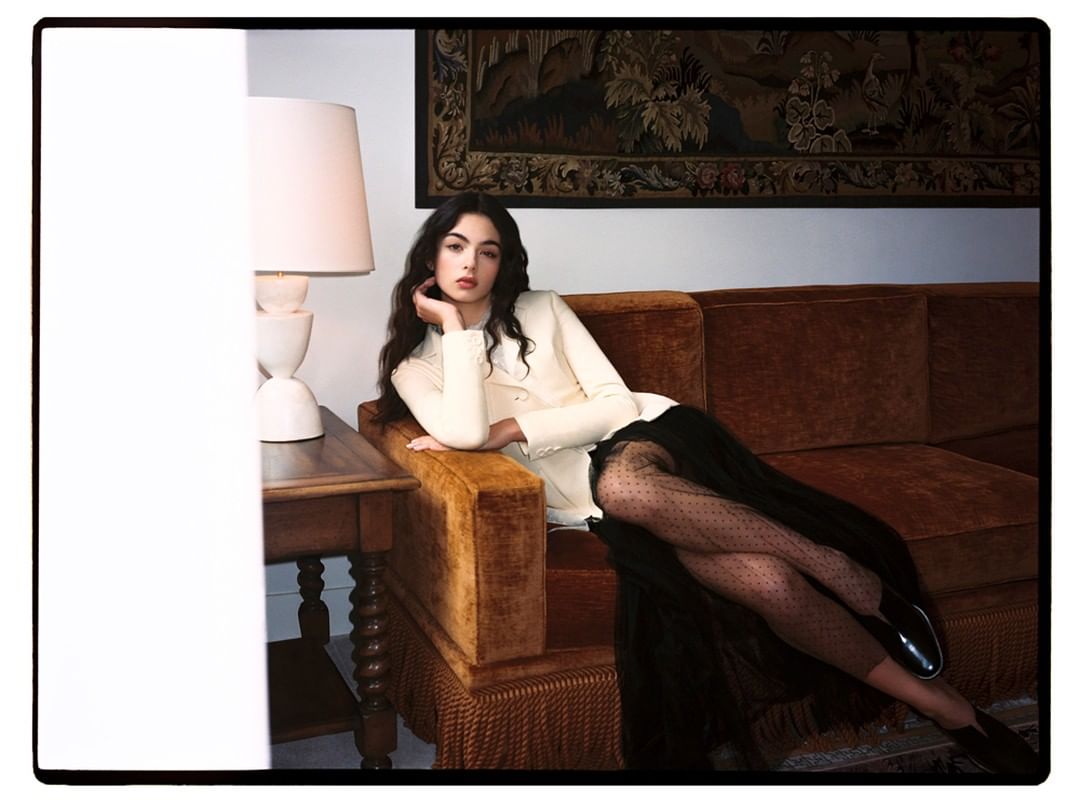 Elegant woman poses on a brown velvet couch in cream blazer and black skirt in an indoor setting with warm, inviting lighting.