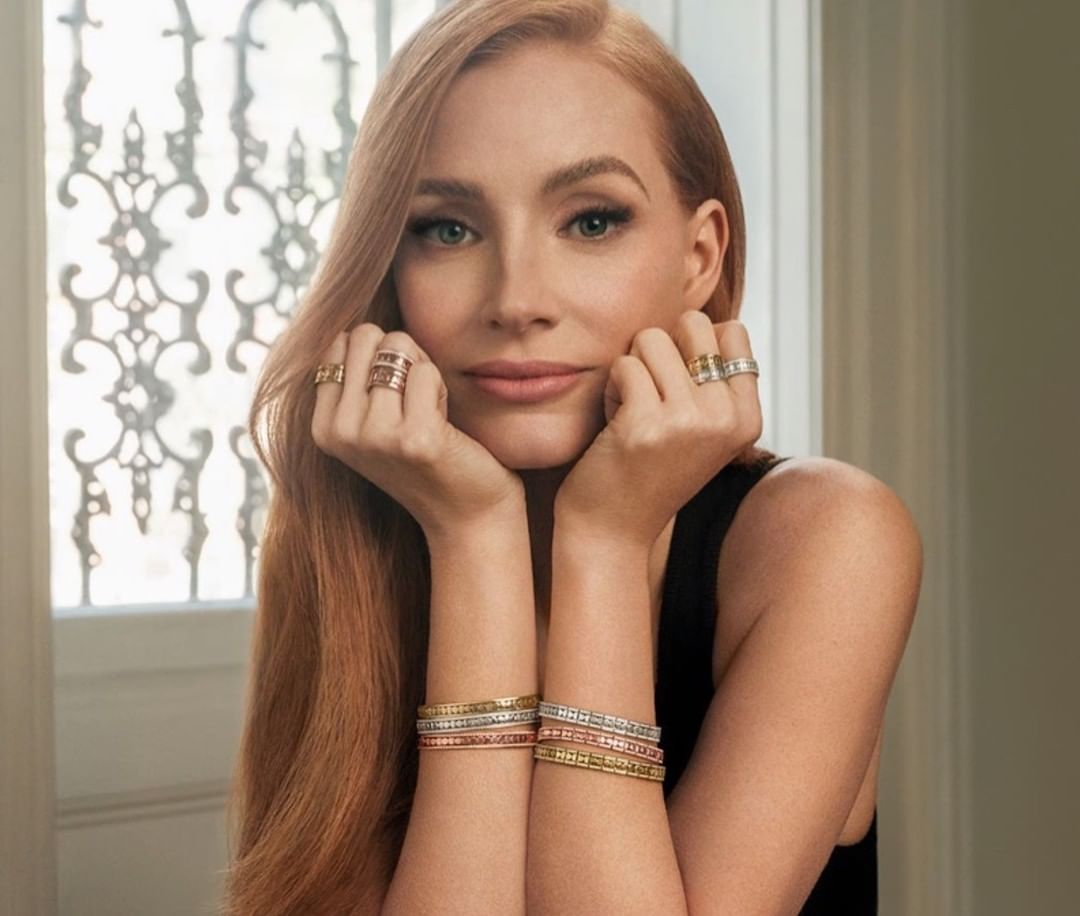 Close-up of a red-haired woman with bracelets and rings, her hands framing her face against a decorative window and soft, diffused lighting.