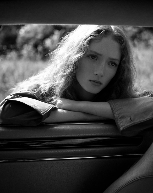 Monochrome portrait of a young woman with curly hair leaning on a car, gazing into the distance in a rural, outdoor environment.