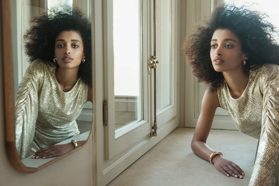 A woman with afro hair, gazing outside, wearing gold sequin dress near window and mirror in an indoor environment with natural light.