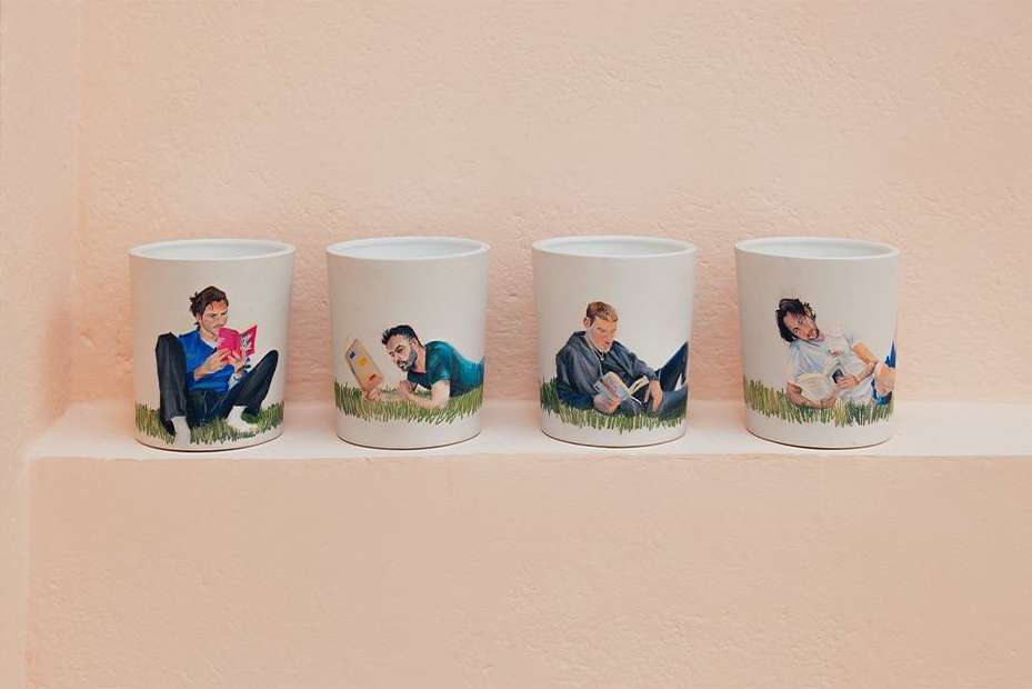 Four cylindrical vessels displaying painted illustrations of men lounging and reading books, arranged on a shelf against a light pink wall.