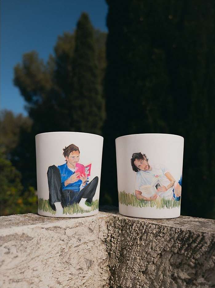 Two white cups feature painted images of men on grass, set against a blurred outdoor backdrop of trees and a clear blue sky.