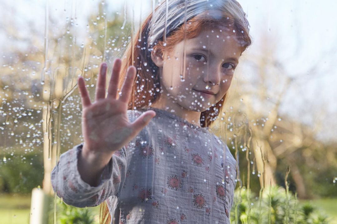 A young red-haired child stands indoors, hand on a rainy window, wearing a floral-patterned dress and headband, with an outdoor background.