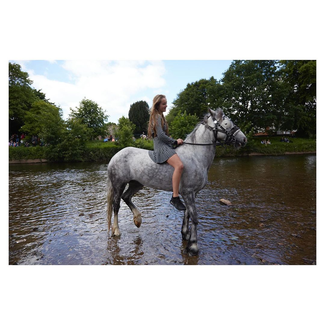 Young woman in a dress riding a gray horse through a shallow river on a sunny day with green trees and blue sky in the background.