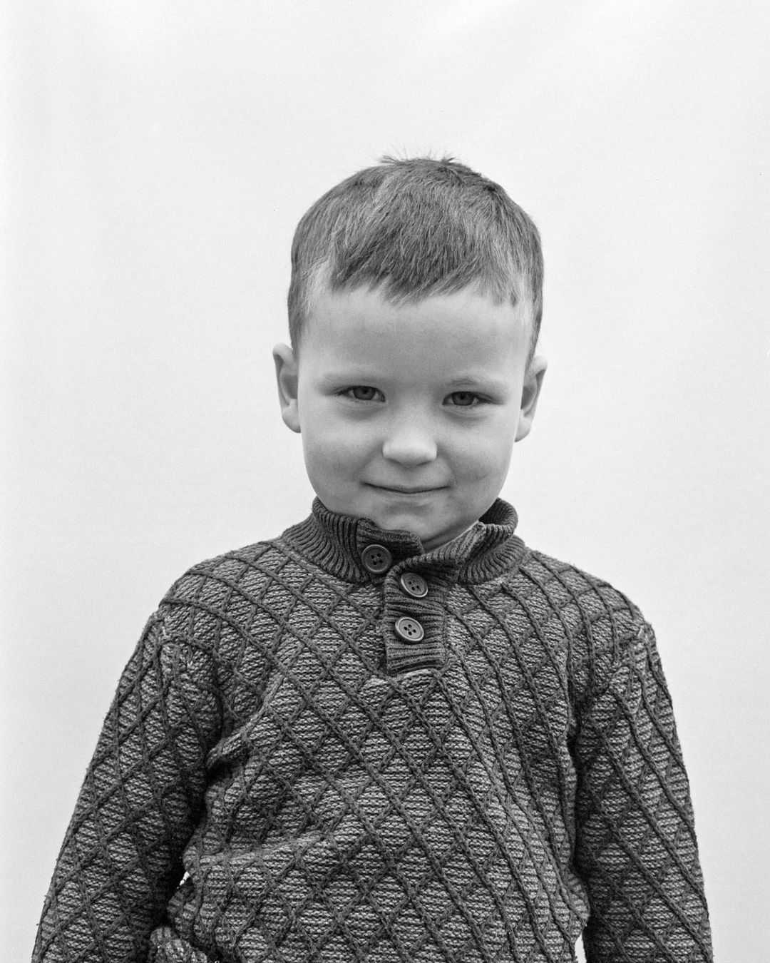 Portrait of a young boy with short brown hair wearing a patterned sweater, set against a plain white background in a studio setting.