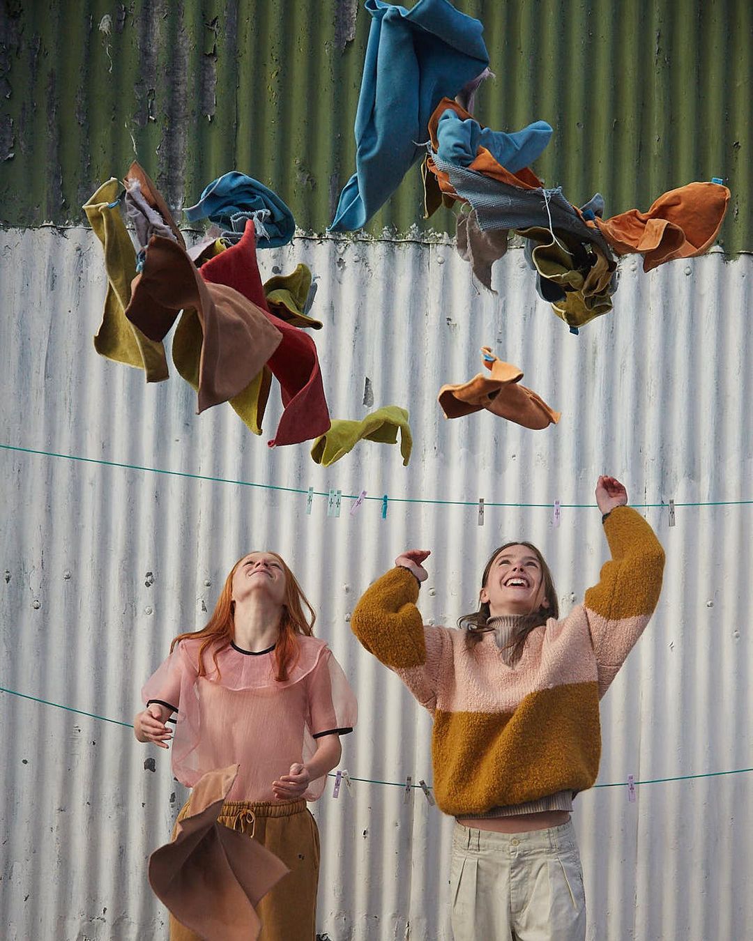 Two smiling women outdoors playfully toss colorful laundry in the air against a textured wall, capturing a lighthearted fashion moment.