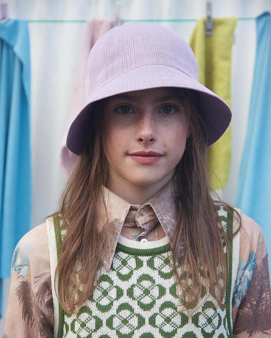 Close-up of a young girl wearing a lilac bucket hat and green patterned vest, posed in front of hanging pastel-colored clothes.