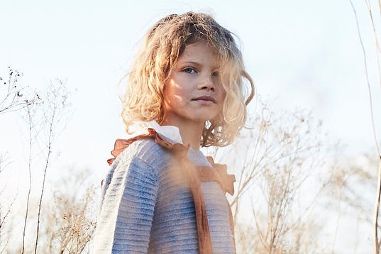 Portrait of a young girl with blonde curly hair wearing a ruffled sweater outdoors in bright sunlight against a soft-focus background.