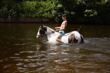A shirtless boy riding a piebald horse through a shallow river, captured by photographer Julia Bostock.
