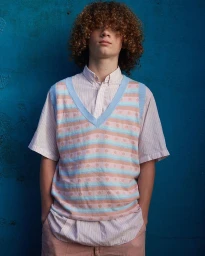 Young man with curly hair wearing a striped sweater vest over a striped button-down shirt against a blue wall.