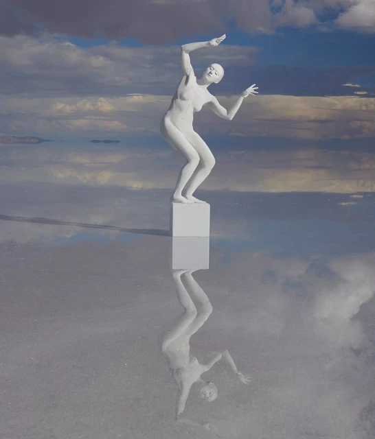 Abstract surreal image of a female figure standing on a cube, mirrored in a shallow body of water under a cloudy sky. 