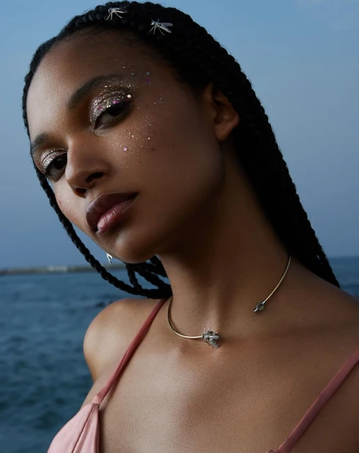 Portrait of an adult woman with dark braided hair, sparkling eye makeup, and a gold choker necklace with a blurred ocean backdrop.
