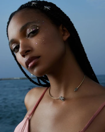 Portrait of an adult woman with dark braided hair, sparkling eye makeup, and a gold choker necklace with a blurred ocean backdrop.