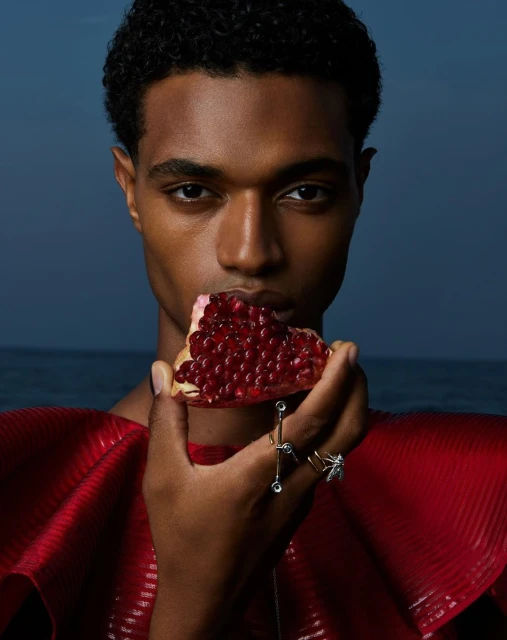 Close-up shot of a handsome dark-skinned man in a red top, biting into a slice of pomegranate with ocean visible in the background.
