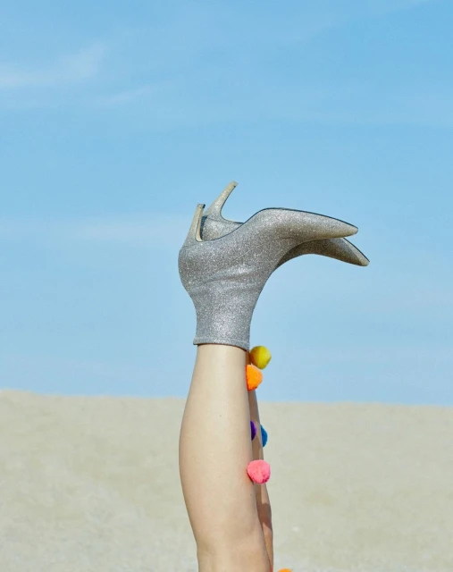 Silver glitter high heel shoe on a woman's leg with colourful pom poms, set against a beach and sky background.