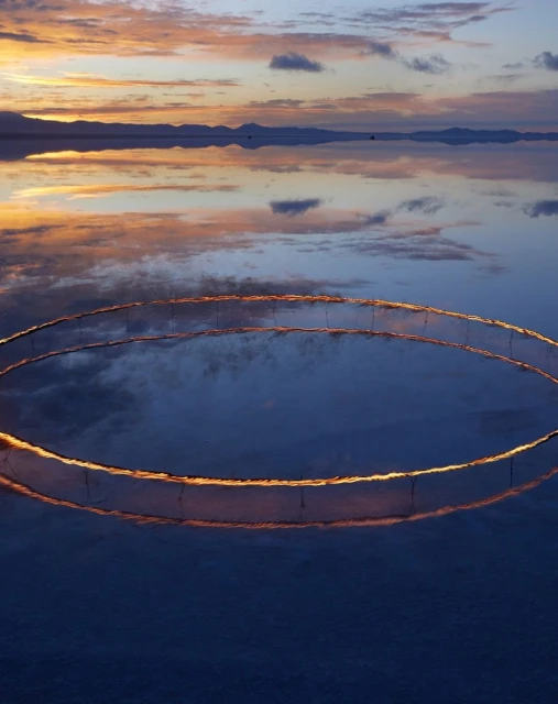 Scenic view of a tranquil body of water reflecting the sky with a vibrant sunrise, featuring a circular installation in the foreground.