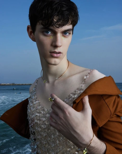 Adult male model poses outdoors wearing a sheer beaded top, gold necklace, ring, and a brown jacket with an ocean backdrop.